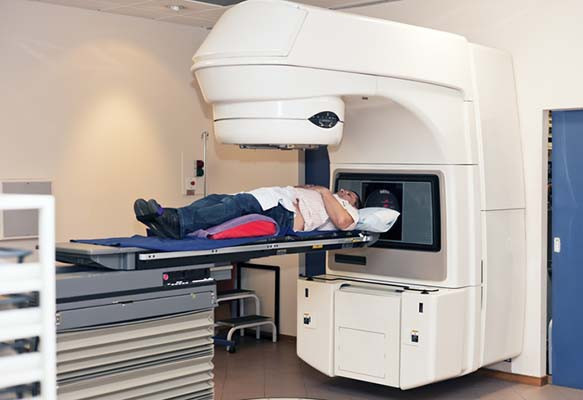 photo of a patient undergoing radiation therapy treatment for cancer; he is seen lying on his back on the bed of a machine with a large white armature hovering above him